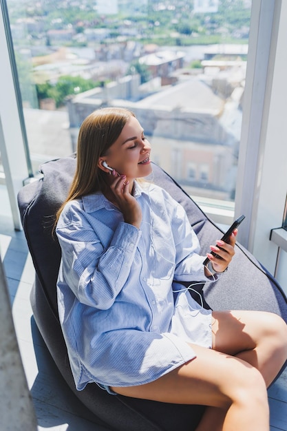 Młoda Kobieta Siedzi Na Fotelu Słuchając Muzyki Na Słuchawkach Za Pomocą Telefonu ładna Kobieta Relaksuje Się Po Dniu Pracy Relaksując Się W Przestrzeni Coworkingowej Z Dużymi Oknami