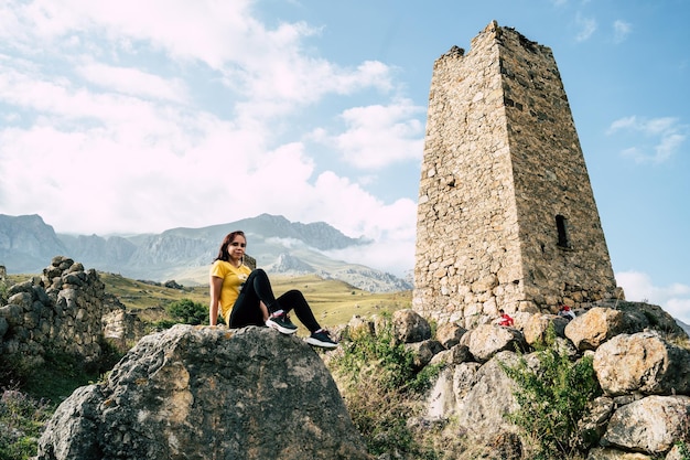 Młoda kobieta siedzi na kamieniu w pobliżu starych wież na tle potężnych gór spowitych chmurami Turystka odpoczywa obok zniszczonych historycznych budynków po aktywnym trekkingu w górach