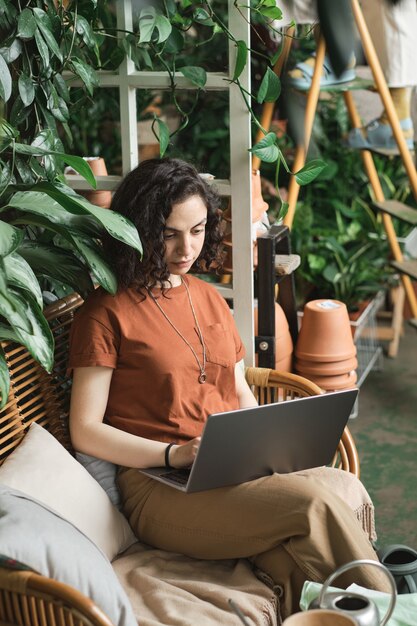 Młoda kobieta siedzi na kanapie i pracuje online na laptopie wśród zielonych roślin w ogrodzie