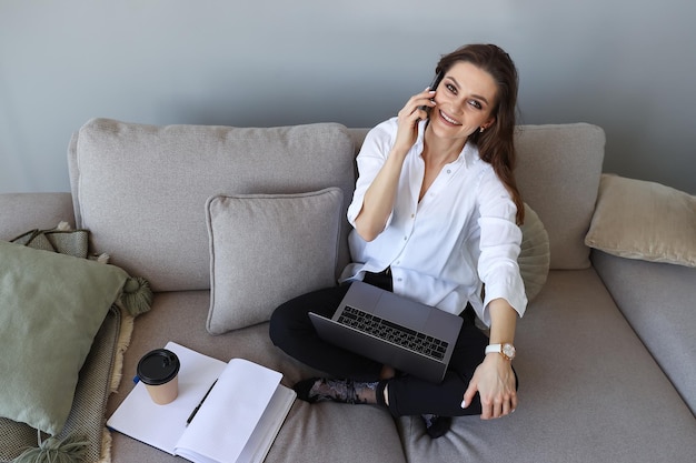 Młoda kobieta siedzi na kanapie i rozmawia przez telefon w domu Praca zdalna