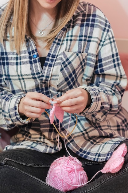 młoda kobieta siedzi na kanapie w domu i robi na drutach różowe serca z przędzy na drutach