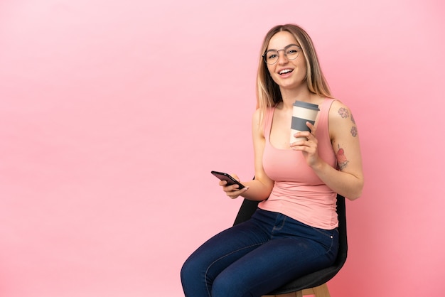 Młoda kobieta siedzi na krześle na różowym tle, trzymając kawę na wynos i telefon komórkowy