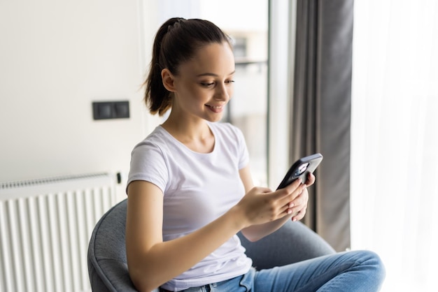 Młoda kobieta siedzi na krześle, używa smartfona, nosi ubrania w stylu casual w domu w pomieszczeniu