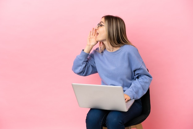 Młoda kobieta siedzi na krześle z laptopem na odosobnionym różowym tle, krzycząc z szeroko otwartymi ustami na boki