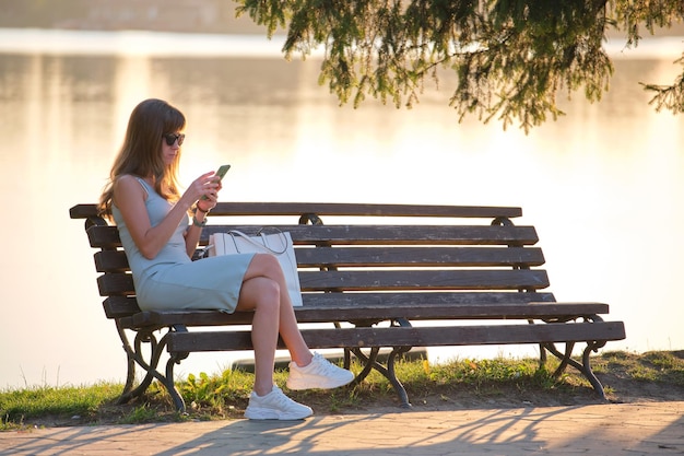 Młoda kobieta siedzi na ławce w parku, przeglądając jej telefon na zewnątrz w ciepły letni wieczór. Koncepcja komunikacji i połączenia mobilnego.