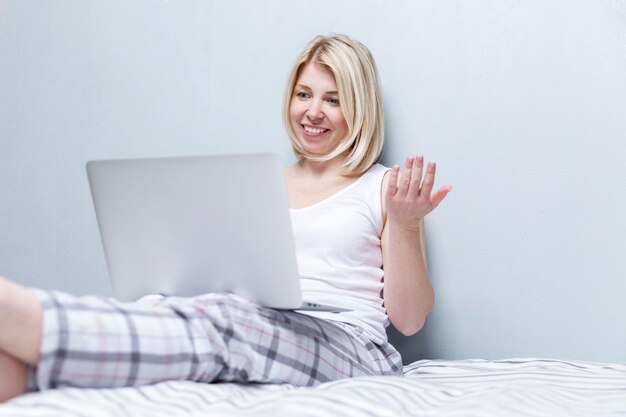 Młoda kobieta siedzi na łóżku z laptopem i rozmawia online Piękna blondynka w piżamie przy szarej ścianie Praca zdalna i komunikacja w sieciach społecznościowych
