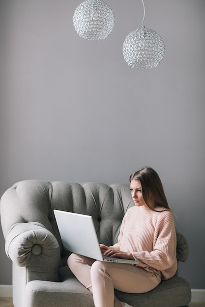 Młoda kobieta siedzi na nowoczesnym fotelu w domu, pracując na laptopie w relaksującej atmosferze.