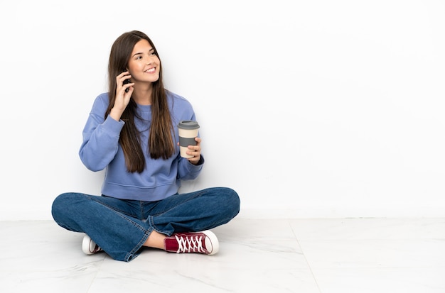 Młoda kobieta siedzi na podłodze trzymając kawę na wynos i telefon komórkowy