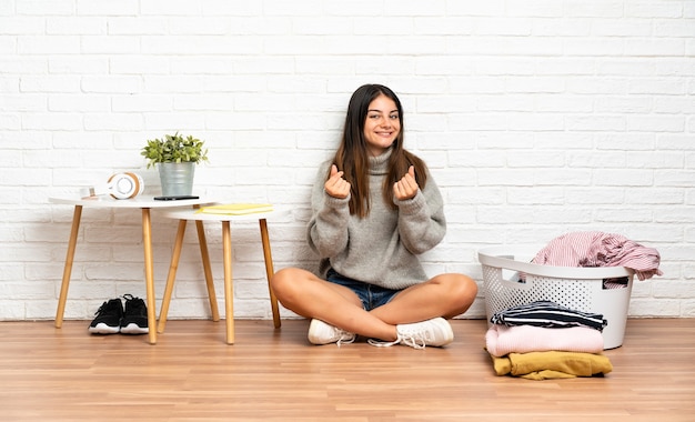 Młoda kobieta siedzi na podłodze w pomieszczeniu z koszem na ubrania, zarabiając gest pieniędzy
