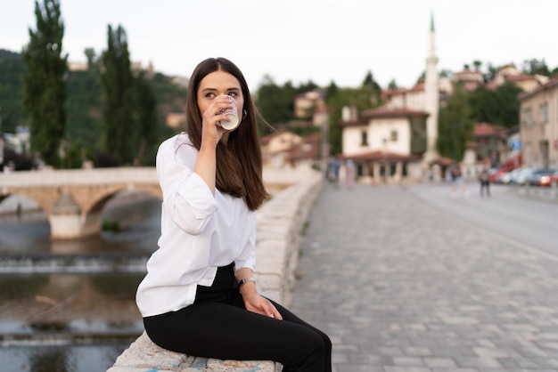 Zdjęcie młoda kobieta siedzi na ścianie w starym mieście i pije kawę.