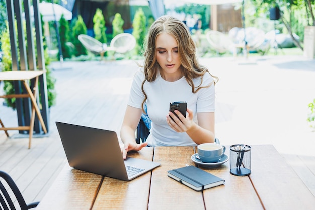 Młoda kobieta siedzi na tarasie kawiarni na ulicy nowoczesnej kawiarni Kobieta siedzi przy stole z telefonem i korzysta z laptopa na świeżym powietrzu Praca zdalna w letniej kawiarni