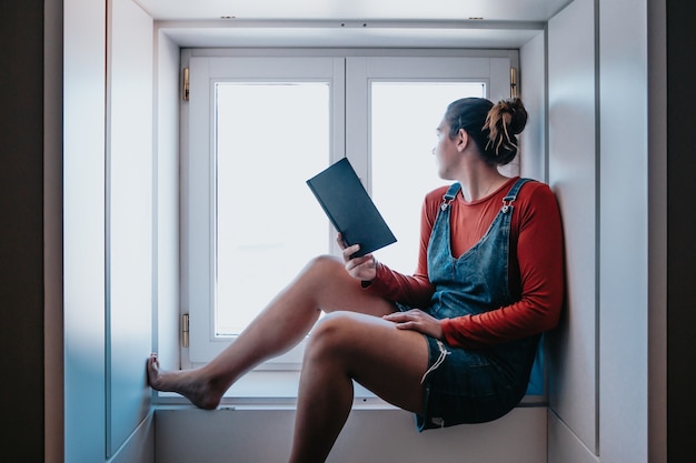 Młoda kobieta siedzi przy oknie i czyta książkę w jasny dzień, koncepcje refleksji i samoobsługi, kopia przestrzeń