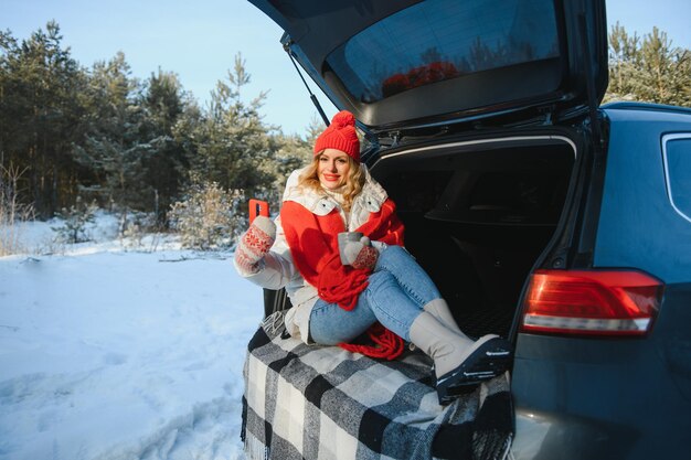 Młoda kobieta siedzi w bagażniku samochodu, pijąc ciepłą herbatę i robi selfie w zimowy, ośnieżony dzień.