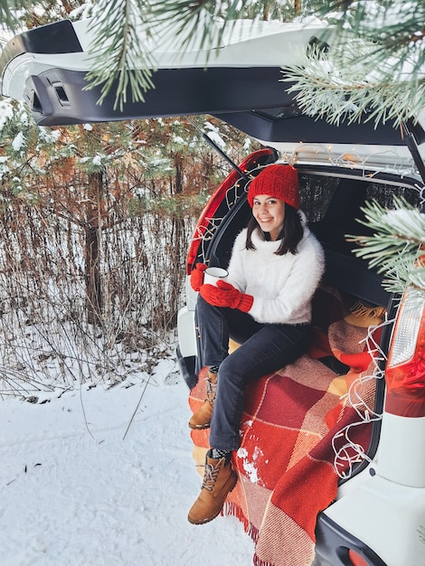Młoda kobieta siedzi w bagażniku samochodu, pijąc ciepłą herbatę w zimowy, zaśnieżony dzień