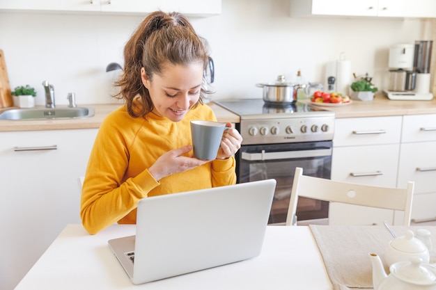 Młoda Kobieta Siedzi W Kuchni W Domu, Używając Na Laptopie