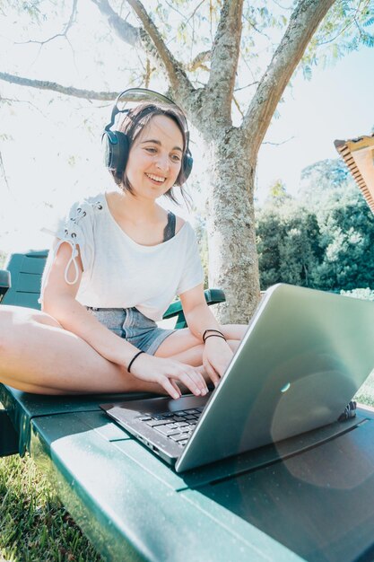 Młoda kobieta siedzi w ogrodzie, uśmiechając się i studiując na uniwersytecie, lubi rekreację na świeżym powietrzu, słuchając muzyki i korzystając z laptopa. Gabinet w domu. Koncepcja życia ludzi. Pisanie na komputerze