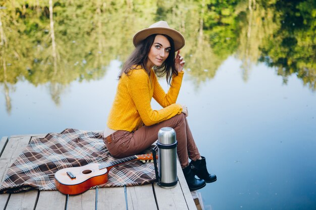 Młoda kobieta siedzi z gitarą na moście nad jeziorem z jesiennym krajobrazem. Tonowanie.