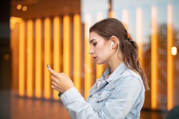 Zdjęcie młoda kobieta słucha muzyki i używa telefonu komórkowego, stojąc przy szklanym oknie