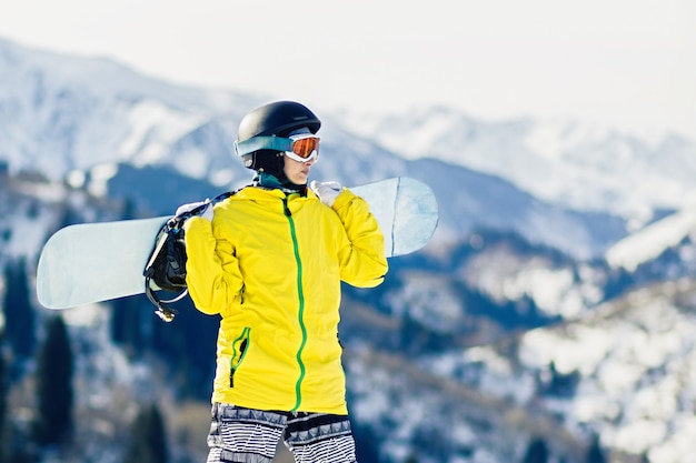 Młoda kobieta snowboardzistka ze snowboardem na ramionach patrzy na stok narciarski, kopia przestrzeń