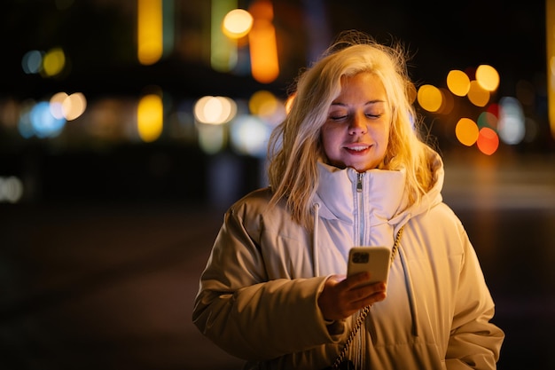 młoda kobieta spaceruje i patrzy na swój telefon komórkowy, który rozświetla jej twarz, uśmiechając się