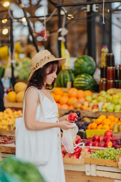 Młoda kobieta stawia owoce i warzywa w bawełnianej torbie na rynku żywności. Ekologiczna torba wielokrotnego użytku na zakupy. Koncepcja zero odpadów.