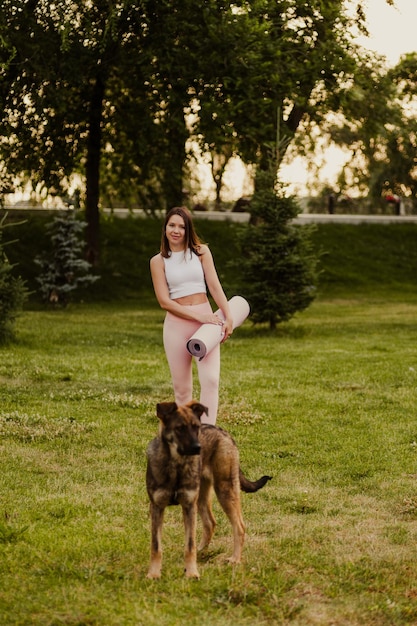 Młoda kobieta stoi na trawie obok psa Atrakcyjna brunetka w sportowych strojach pozuje na tle malowniczych drzew i trzyma w rękach matę do jogi i pilatesu
