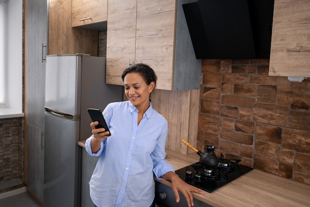 Młoda kobieta, stojąc w kuchni, czyta wiadomości tekstowe na smartfonie.