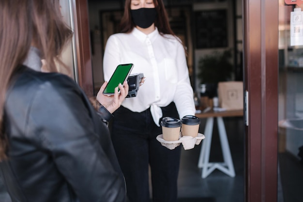 Młoda kobieta stojąca na zewnątrz w masce i płacąca rachunek za pośrednictwem smartfona przy użyciu technologii NFC w formacie