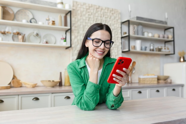 Młoda kobieta stojąca w kuchni w domu, opierając się na stole i sprawdzając czat na telefonie komórkowym