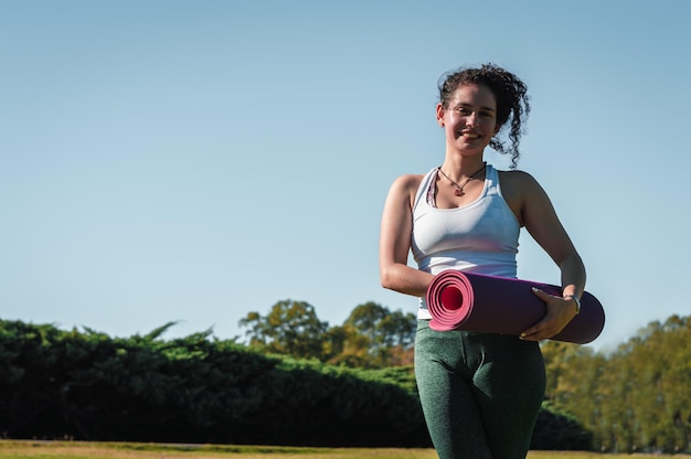 młoda kobieta stojąca w parku uśmiecha się z różową matą do jogi w dłoniach