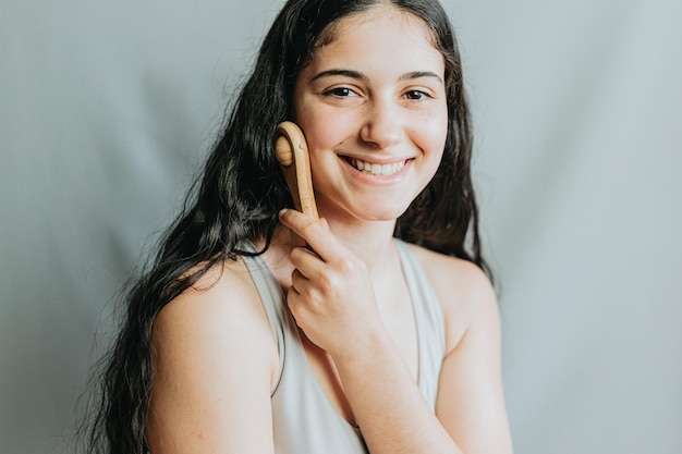 Młoda kobieta stosująca masaż twarzy drewnianą rolką Radosny dzień samoopieki Wywieranie nacisku w celu poprawy krążenia Nowe metody pielęgnacji skóry i codziennych rytuałówPortret w studio z szarym tłem
