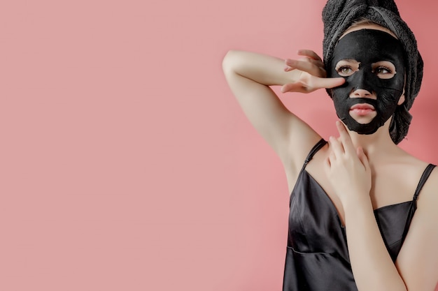 Młoda kobieta stosuje czarną tkaninę kosmetyczną twarzy maski na różowym tle. Maska peelingująca z węglem drzewnym, zabiegi kosmetyczne w spa, pielęgnacja skóry, kosmetologia. Ścieśniać