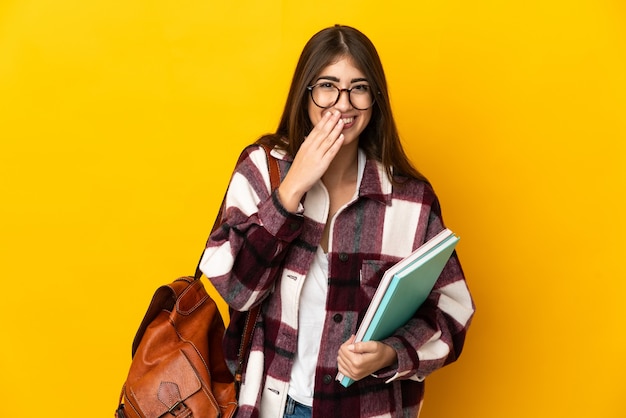 Młoda kobieta student na białym tle na żółtym tle szczęśliwa i uśmiechnięta obejmująca usta ręką