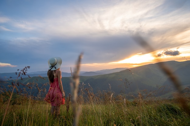 Młoda Kobieta Szczęśliwa Podróżnik W Czerwonej Sukience Stojący Na Trawiastym Zboczu Wzgórza W Wietrzny Wieczór W Górach Latem, Ciesząc Się Widokiem Natury O Zachodzie Słońca.