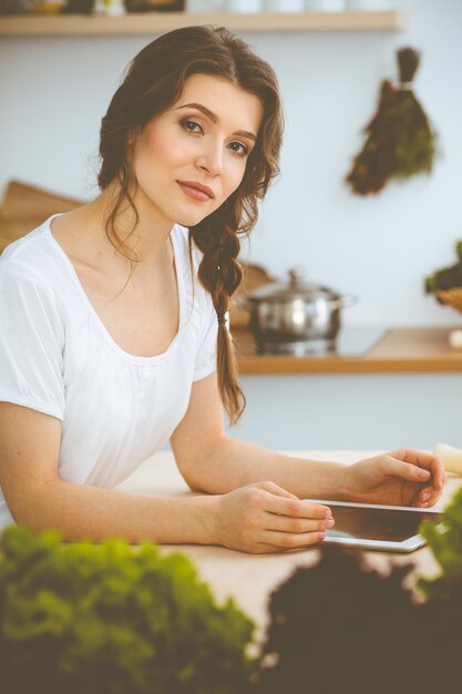 Młoda kobieta szuka nowego przepisu na gotowanie w kuchni. Gospodyni robi zakupy online za pomocą komputera typu tablet i karty kredytowej.