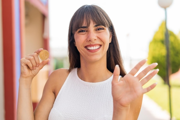 Młoda kobieta trzyma Bitcoina na zewnątrz pozdrawiając ręką z radosnym wyrazem twarzy