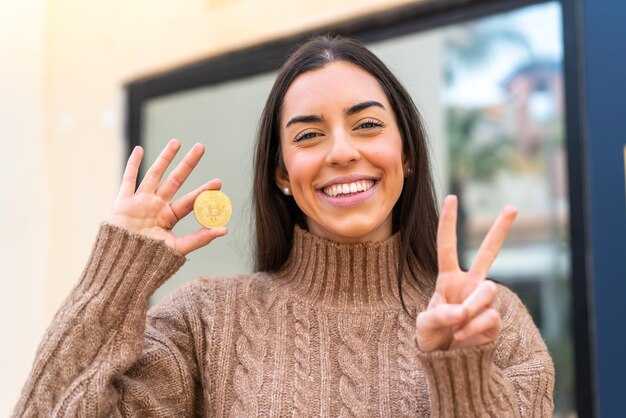 Młoda Kobieta Trzyma Bitcoina Na Zewnątrz, Uśmiechając Się I Pokazując Znak Zwycięstwa
