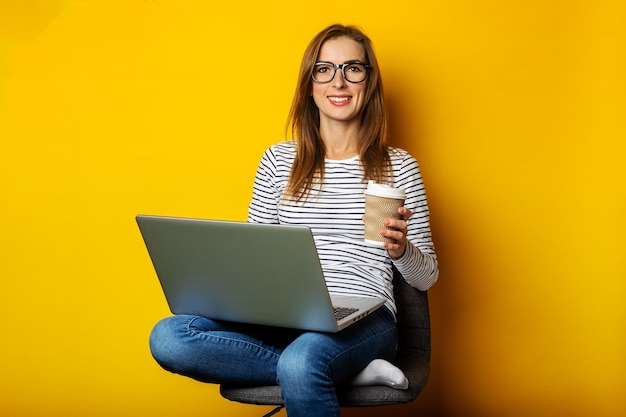 Młoda kobieta trzyma filiżankę kawy, siedząc na krześle, pracując na laptopie na białym tle żółty.