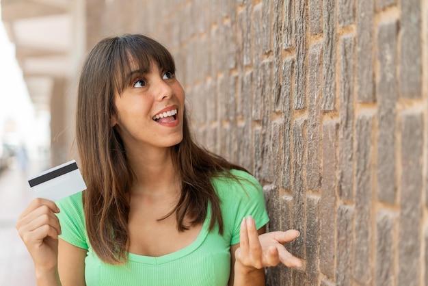 Młoda kobieta trzyma kartę kredytową na zewnątrz z niespodzianką wyraz twarzy