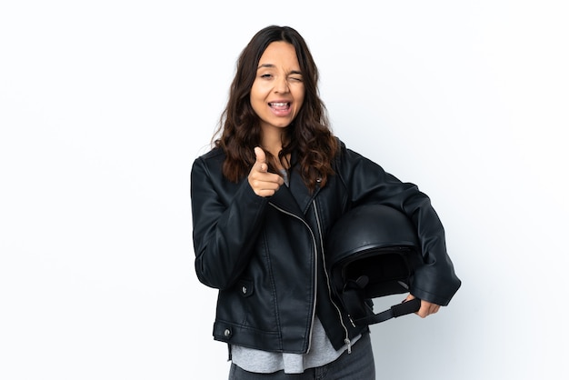 Młoda kobieta trzyma kask motocyklowy na pojedyncze białe tło, wskazując na przód i uśmiechnięte