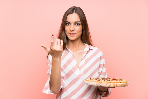 Młoda kobieta trzyma pizzę nad odosobnioną menchii ścianą zaprasza przychodzić z ręką. Cieszę się, że przyszedłeś