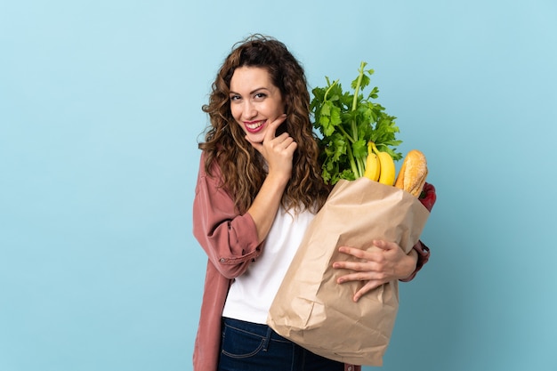 Młoda kobieta trzyma torbę na zakupy spożywcze na białym tle na niebieskim tle szczęśliwa i uśmiechnięta