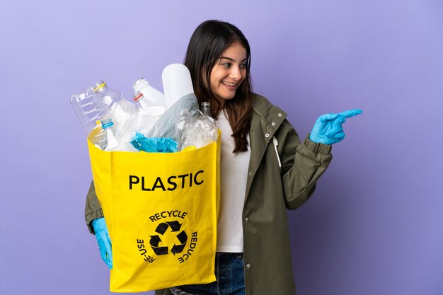 Młoda kobieta trzyma torbę pełną plastikowych butelek do recyklingu na białym tle na fioletowej ścianie, wskazując palcem w bok i prezentuje produkt