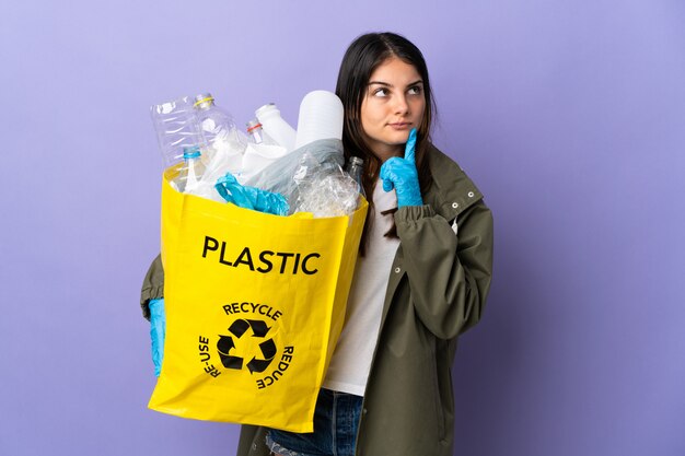 Młoda kobieta trzyma torbę pełną plastikowych butelek do recyklingu na białym tle na fioletowo, mając wątpliwości, patrząc w górę