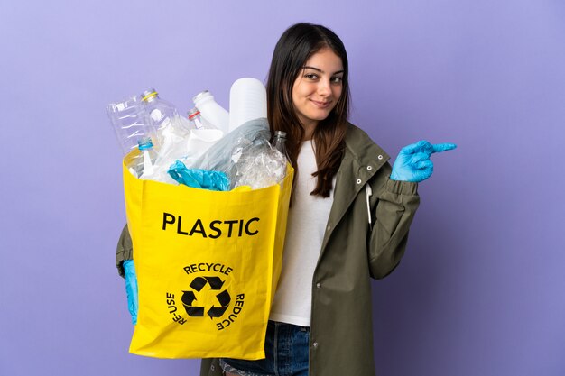 Młoda kobieta trzyma torbę pełną plastikowych butelek do recyklingu na białym tle na fioletowy palec wskazujący na bok
