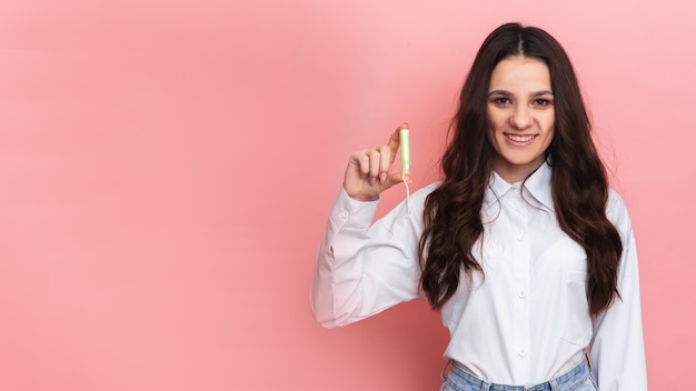 Młoda kobieta trzyma w dłoniach wacik menstruacyjny z aplikatorem dla łatwego użycia. Różowe tło. Miejsce na tekst