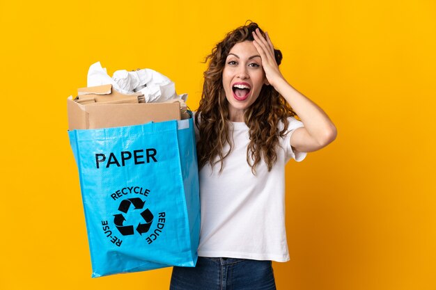 Młoda kobieta trzyma worek recyklingu pełen papieru do recyklingu na białym tle na żółtej ścianie z wyrazem zaskoczenia