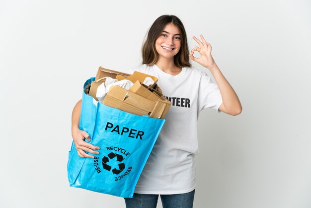 Młoda kobieta trzyma worek recyklingu pełnego papieru do recyklingu na białym, pokazując znak ok palcami