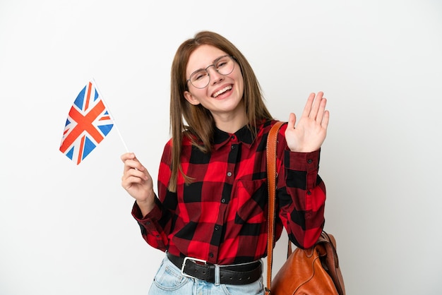 Młoda kobieta trzymająca flagę Wielkiej Brytanii na białym tle na niebieskim tle, pozdrawiając ręką ze szczęśliwym wyrazem twarzy