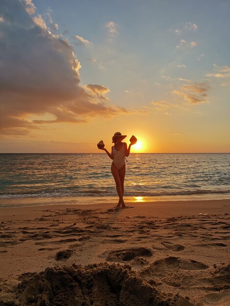 Zdjęcie młoda kobieta trzymająca kokosy stojąca na plaży na tle nieba podczas zachodu słońca